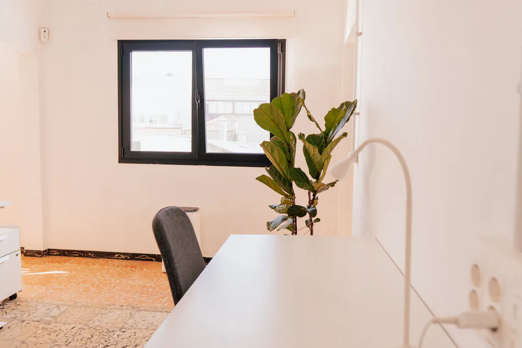 Puesto junto a la ventana con mesa blanca, flexo y una planta.